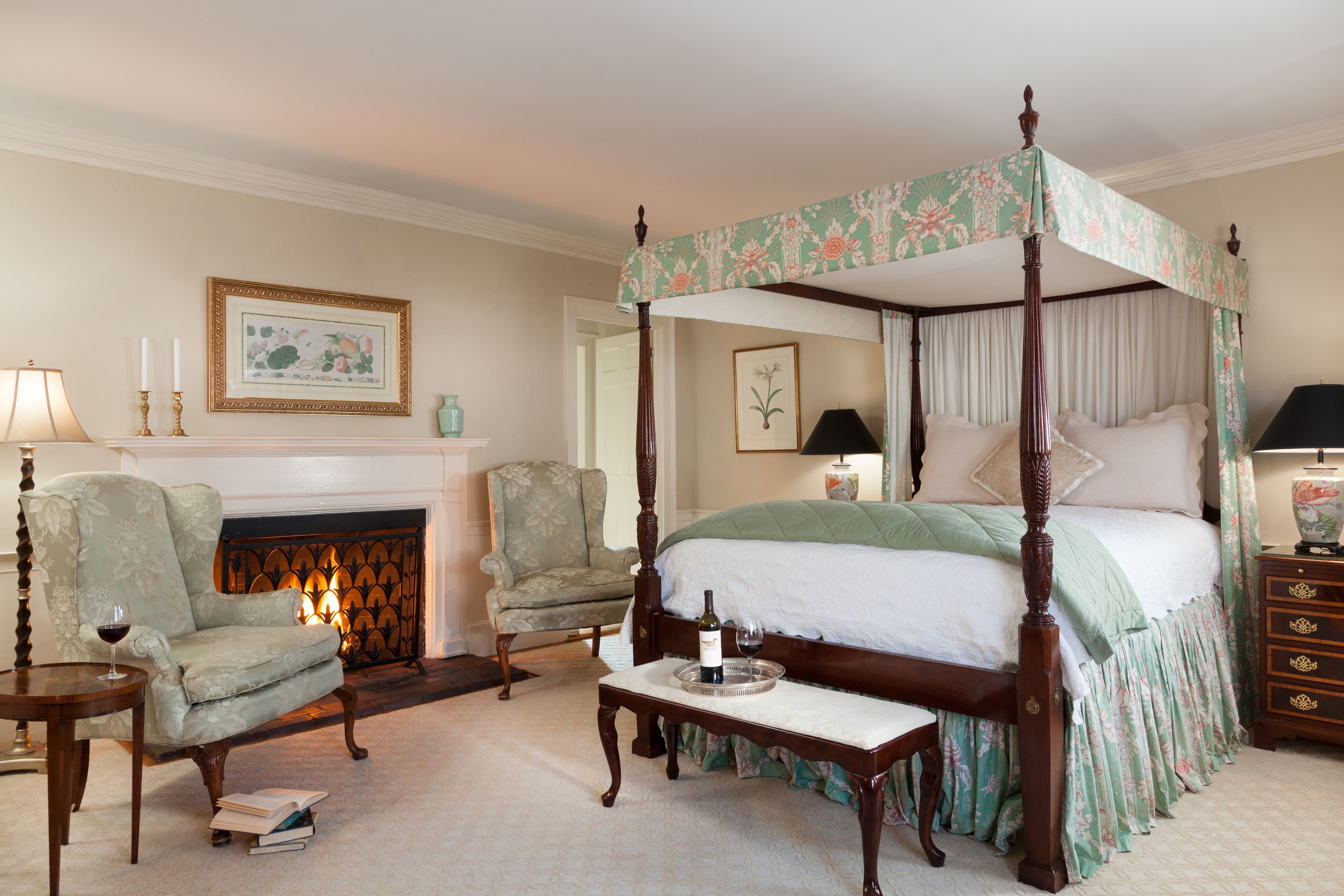 A four-poster bed with a canopy, a fireplace, two armchairs, a bedside table with a tray, and a framed picture on the wall.