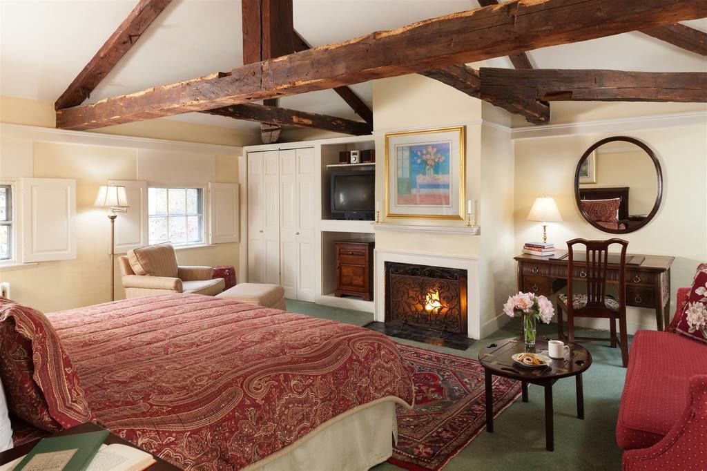 A traditional room with a fireplace, a bed with a red patterned bedspread, a desk with a mirror, and a chair.