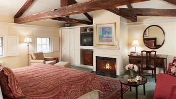A traditional room with a fireplace, a bed with a red patterned bedspread, a desk with a mirror, and a chair.