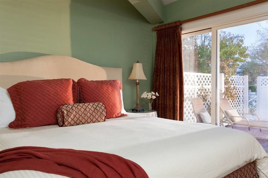 A bedroom with a large bed, a nightstand with a lamp, and a view of a patio with chairs.