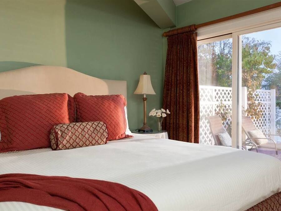 A bedroom with a large bed, a nightstand with a lamp, and a view of a patio with chairs.