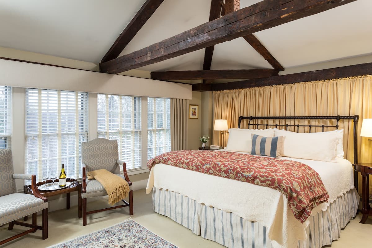 A bedroom with a large bed, a wooden headboard, a window with blinds, a chair, and a small table with a bottle and glasses.
