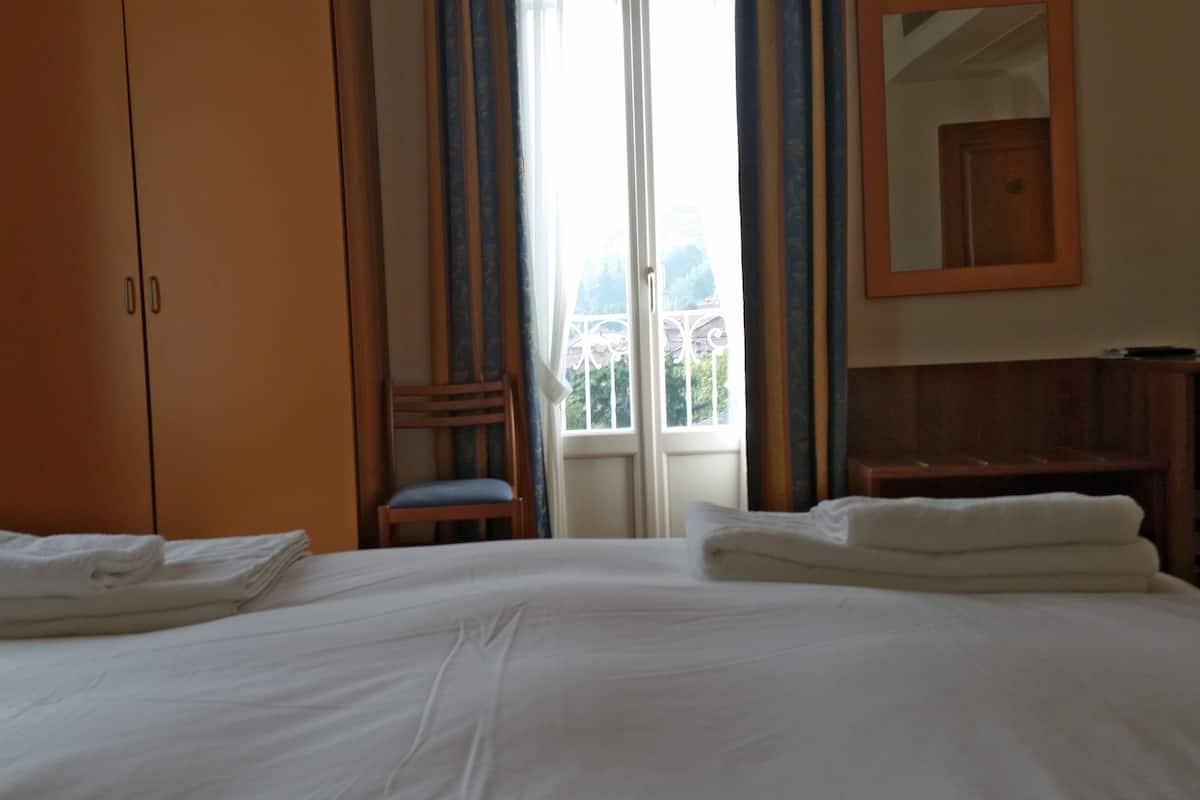 A hotel room with a large bed, a wooden wardrobe, a chair, and a view of the outdoors through a window.