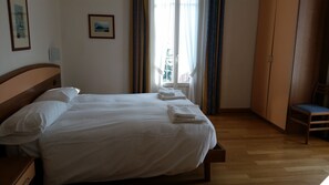 A neatly made bed with white linens and towels, a wooden headboard, a wooden nightstand, a chair, and a window with curtains.