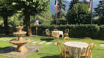 Ein Garten mit einer Steinfontäne, gelben Korbstühlen und einem Tisch mit einer Blumen-Tischdecke.