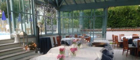 An outdoor dining area with tables set for a meal, covered with white tablecloths and adorned with pink flowers in glass vases.