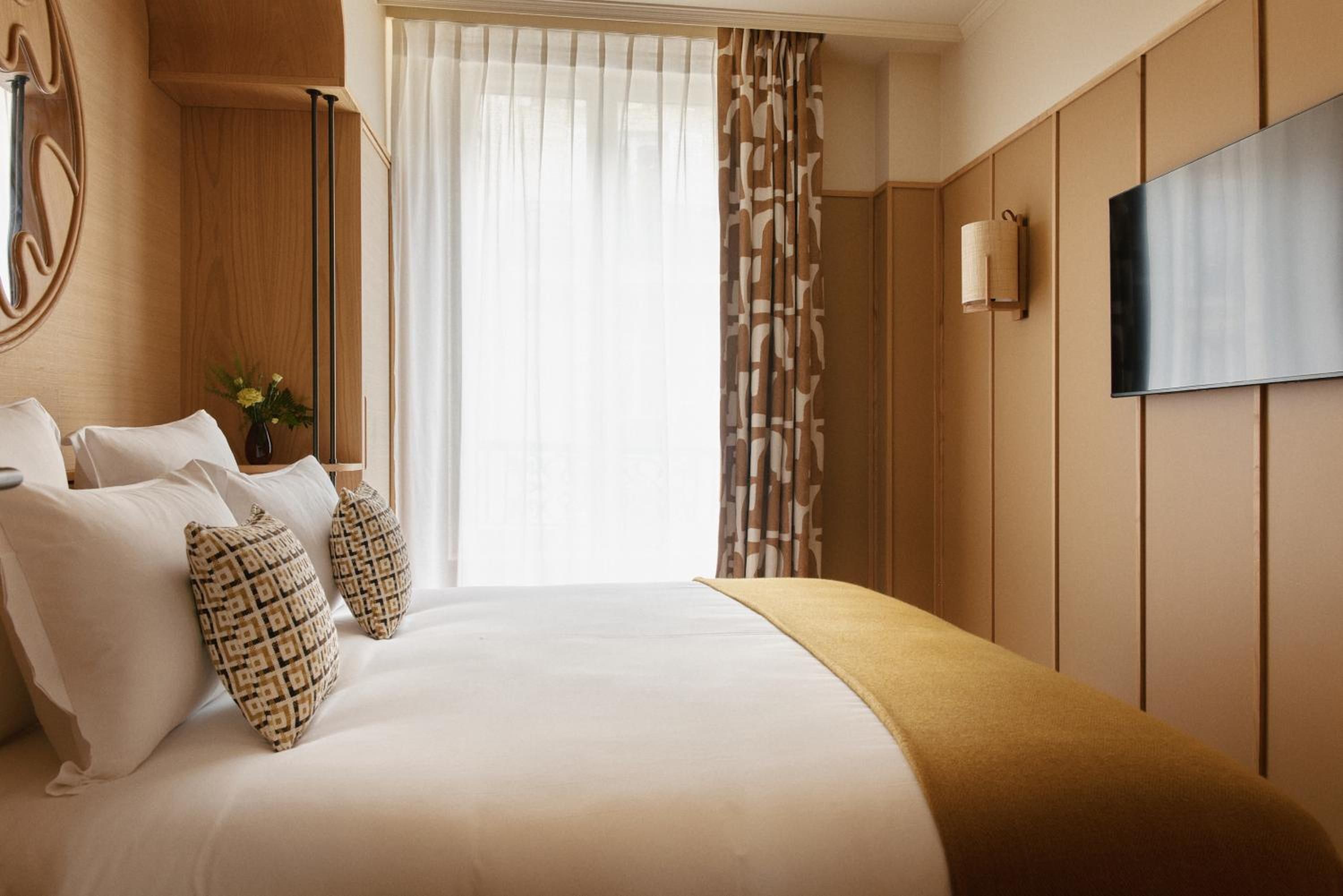 A hotel room with a large bed, a flat-screen TV mounted on the wall, and a window with sheer curtains.