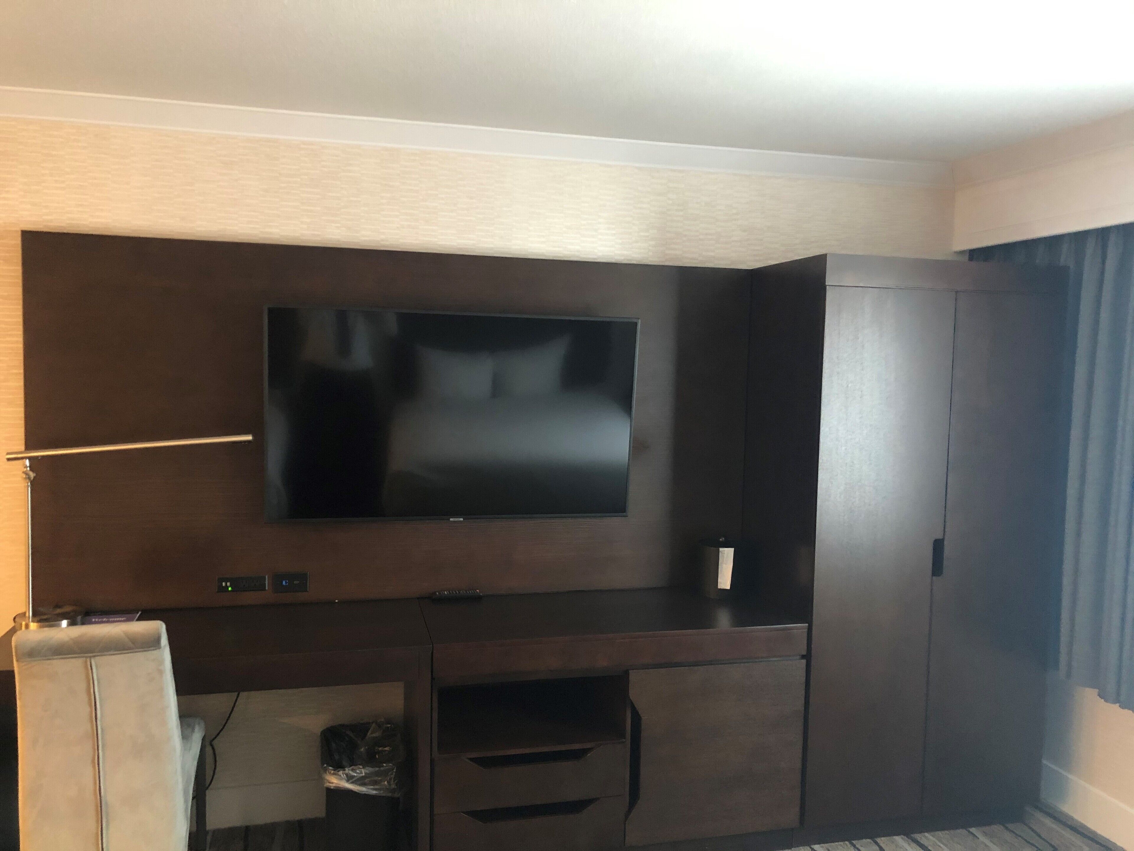 A hotel room with a large flat-screen TV mounted on a dark wooden wall, a small desk with drawers, and a wardrobe.