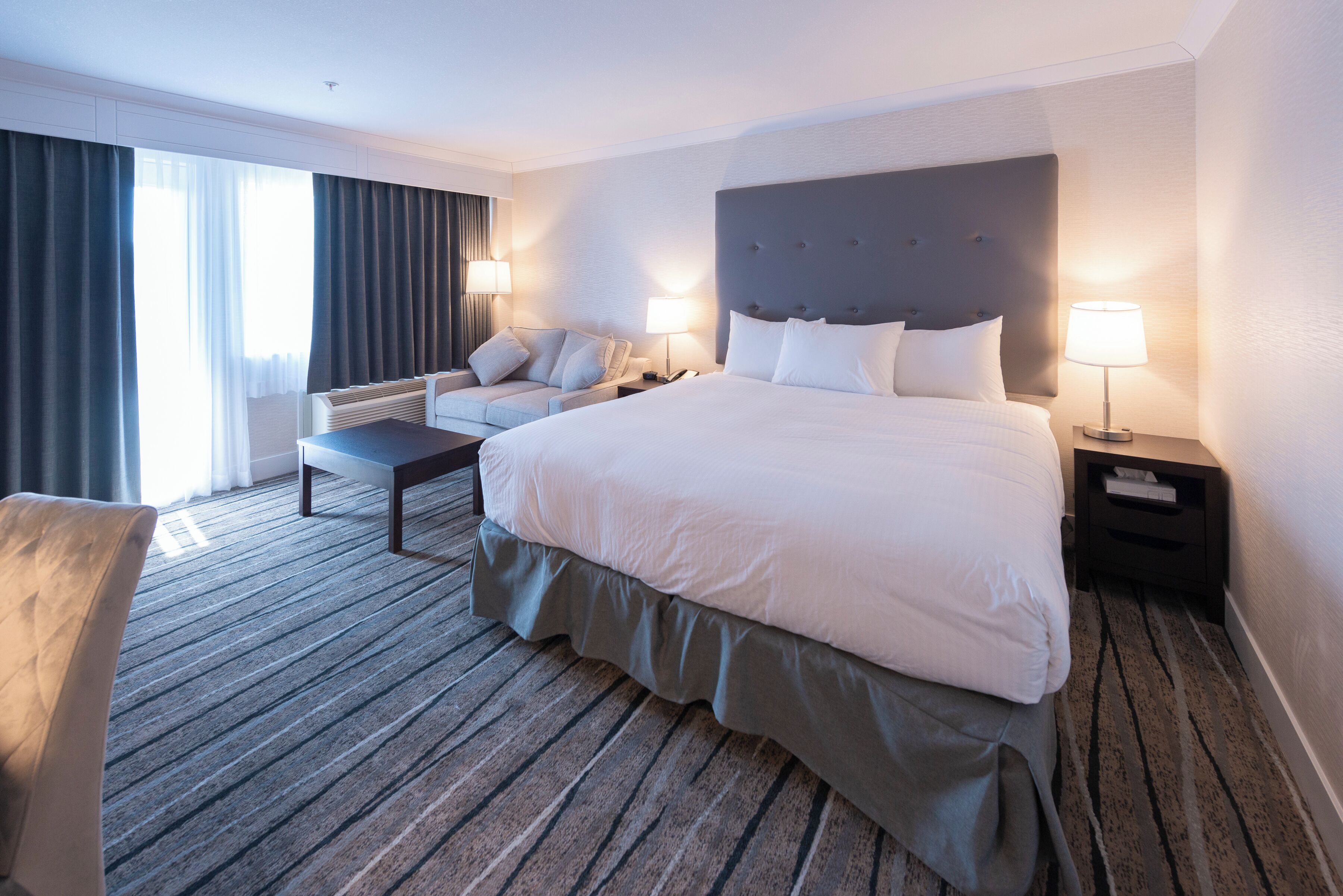 A hotel room with a large bed, a sofa, a desk, and a chair. There are two bedside tables with lamps.