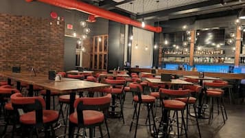 A restaurant with multiple tables and red chairs, a bar area with bottles, and a brick wall.