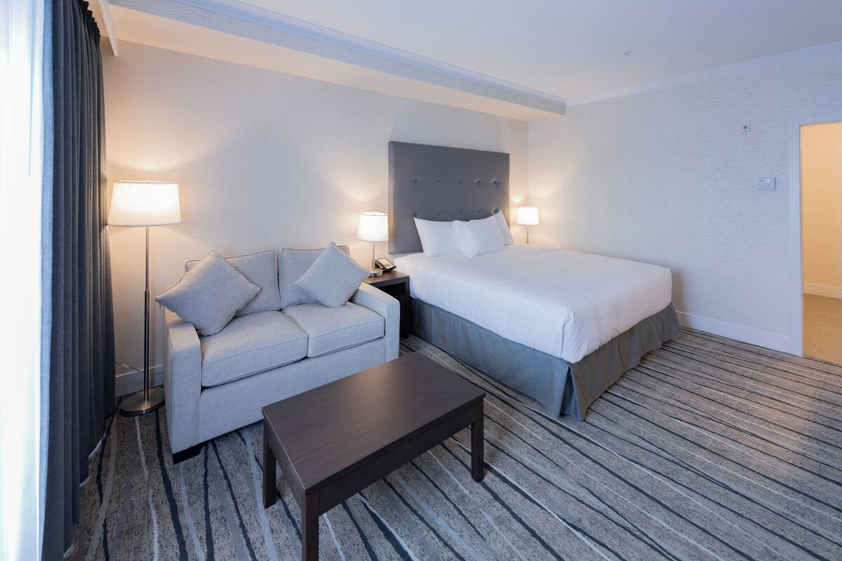 A hotel room with a bed, a sofa, a coffee table, a floor lamp, and a bedside table with a lamp.