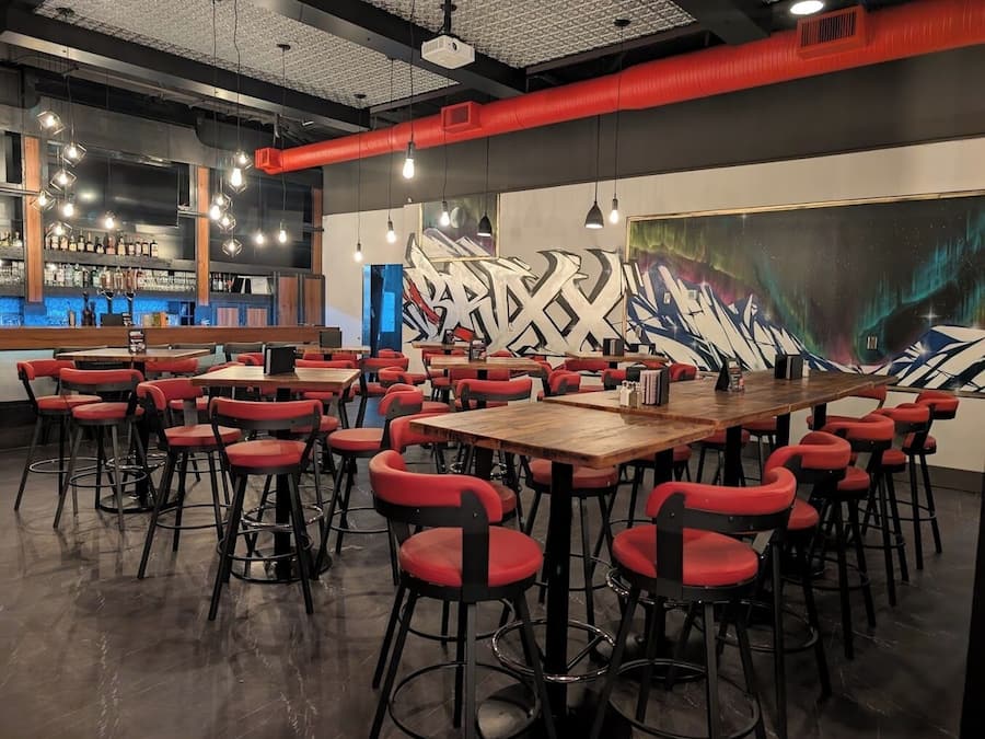 A restaurant with multiple tables and red chairs, a bar area with bottles, and a large mural on the wall.