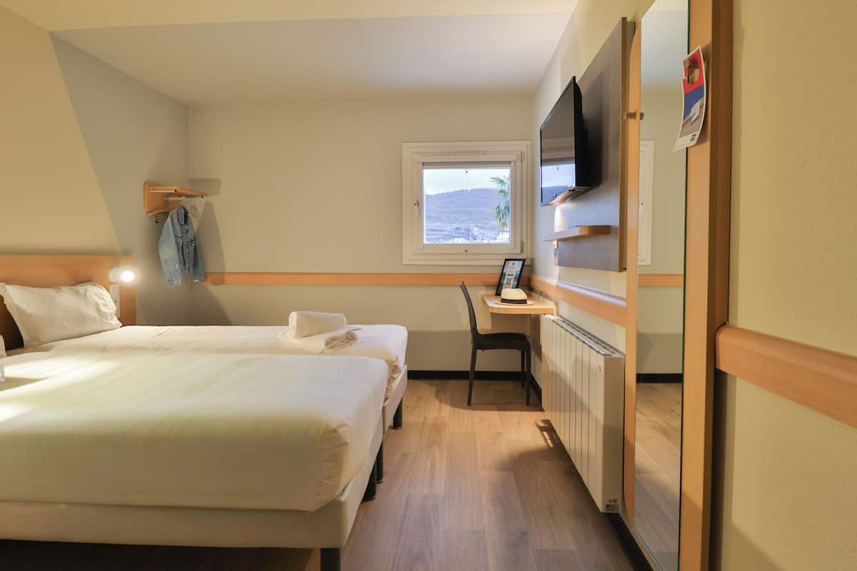 Une chambre d’hôtel équipée d’un lit, d’un bureau, d’une chaise, d’une tél évision et d’une fenêtre donnant sur des collines.