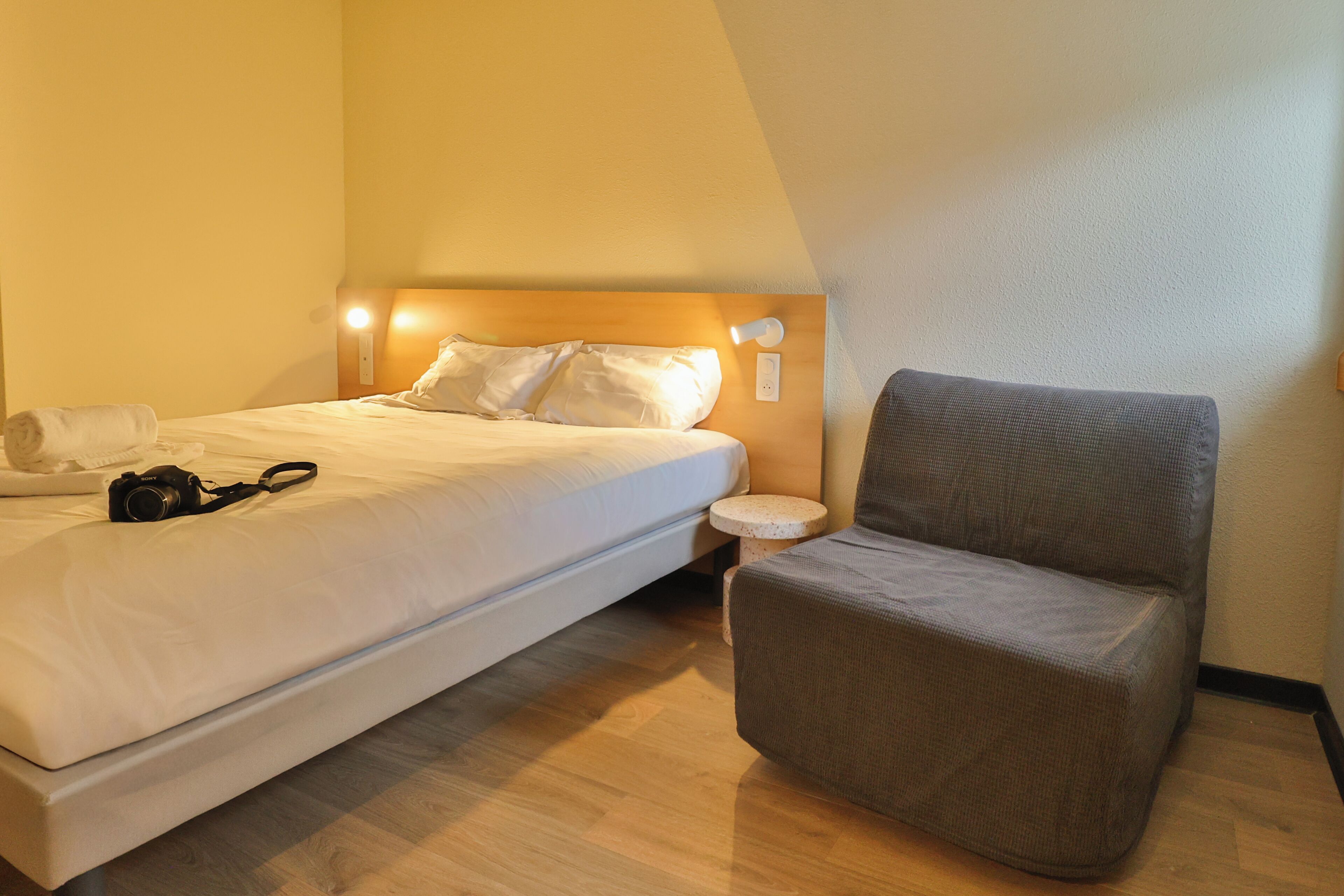 A hotel room with a bed, a grey armchair, a small round table, and a wall-mounted light fixture.