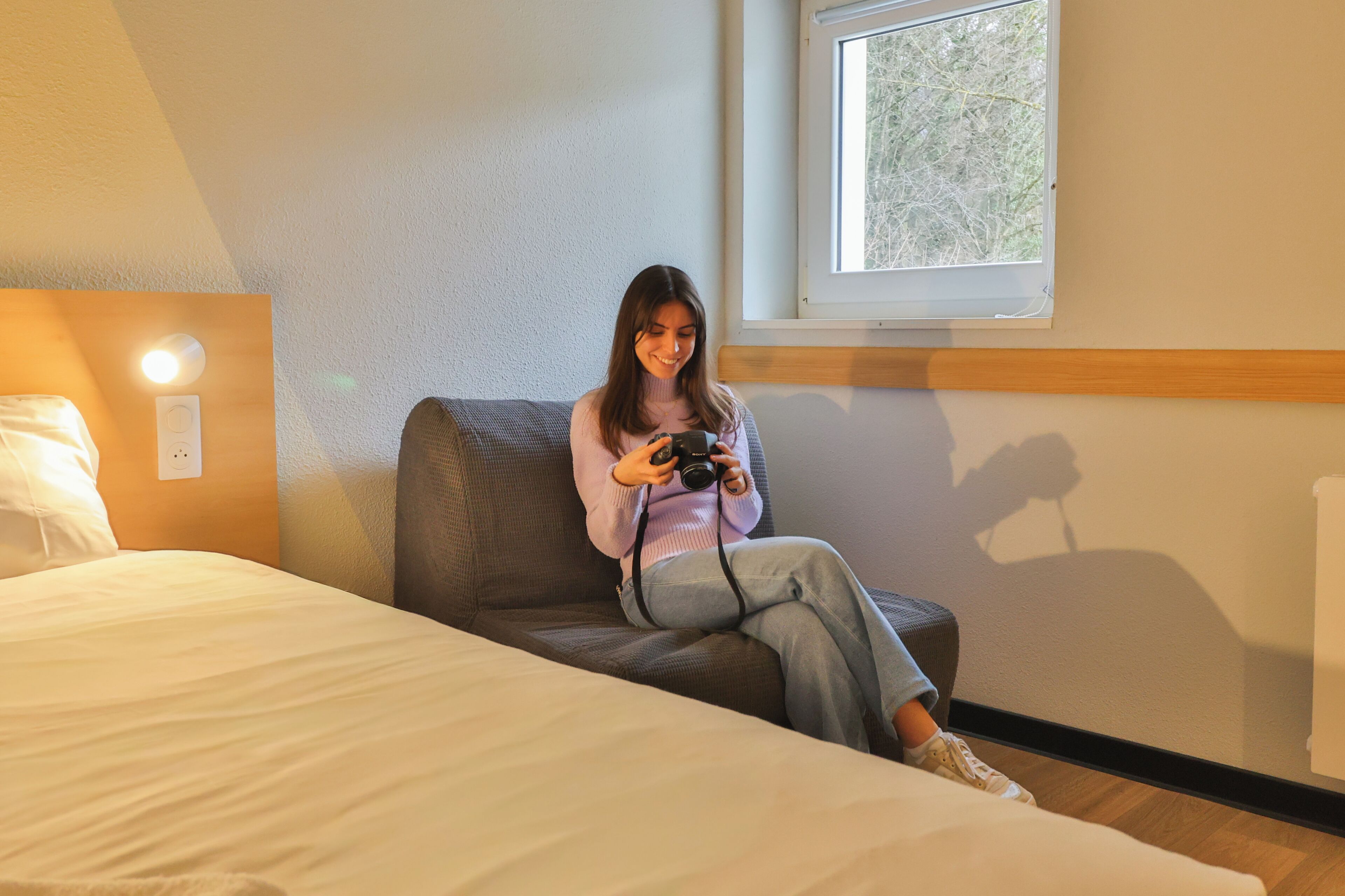 A person sitting on a bed in a room with a window, a wall socket, and a wooden headboard.