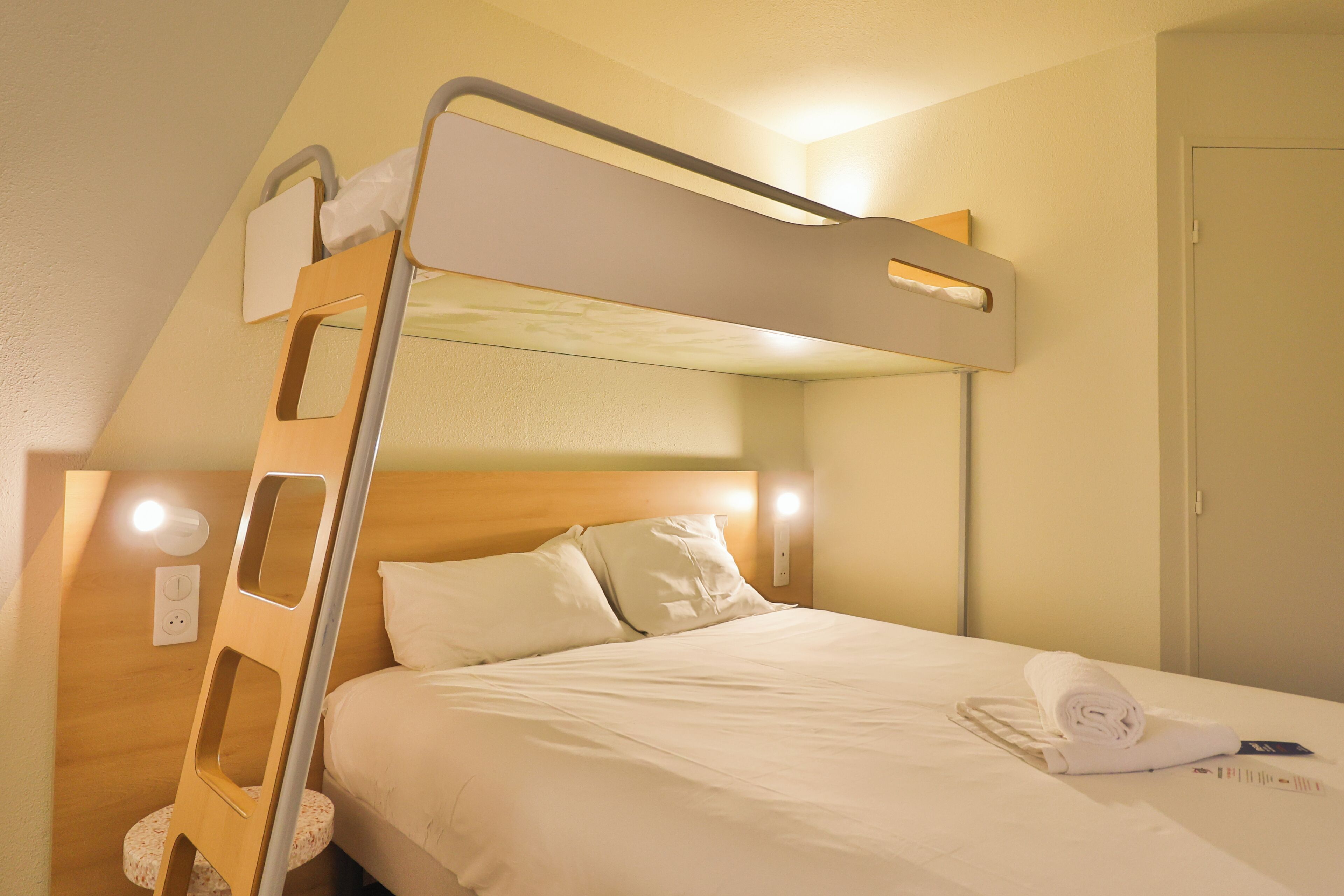 Een stapelbed met een houten trapje, een nachtkastje met een lamp, en een netjes opgemaakt bed met een opgerolde handdoek.