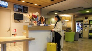 A reception desk with a flat-screen TV, a vase with dried flowers, and a green trash bin. - Hôtel ibis budget Aix-les-Bains Nord (Gresy-sur-Aix)