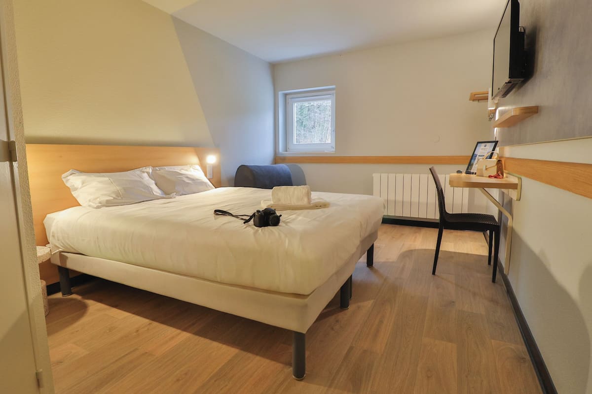 Une chambre d’hôtel comprenant un lit, un bureau avec une chaise, un téléviseur fixé au mur et une fenêtre laissant entrer la lumière naturelle.