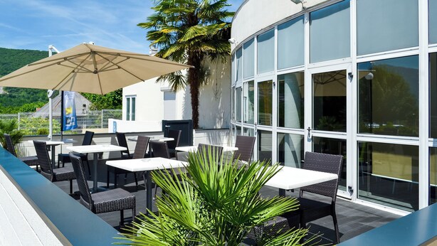An outdoor patio with tables and chairs, a large umbrella, and a palm tree. - Hôtel ibis budget Aix-les-Bains Nord (Gresy-sur-Aix)