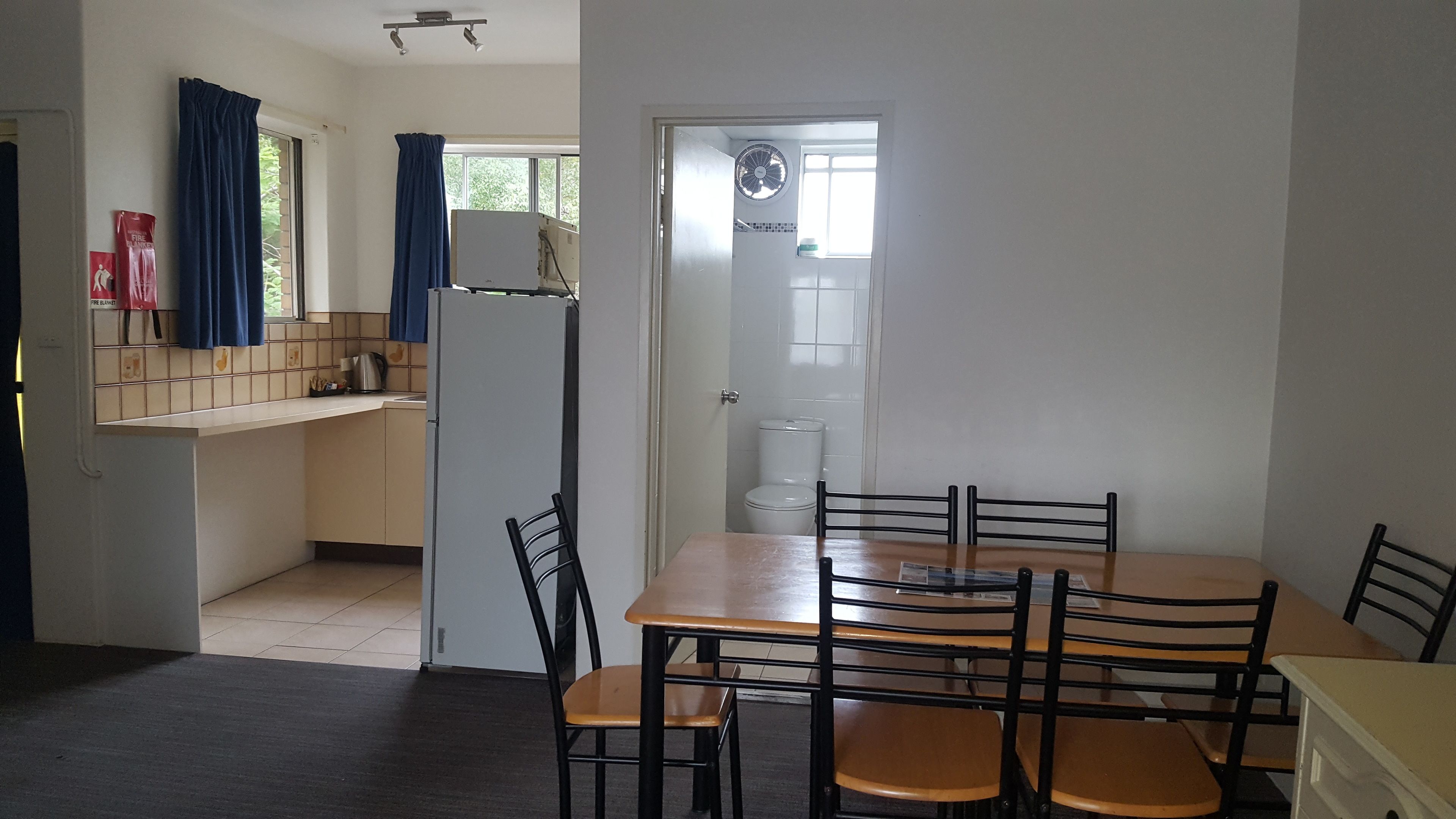 Una cocina compacta con refrigerador, pileta y ventana con cortinas. Hay una zona de comedor con mesa y sillas.