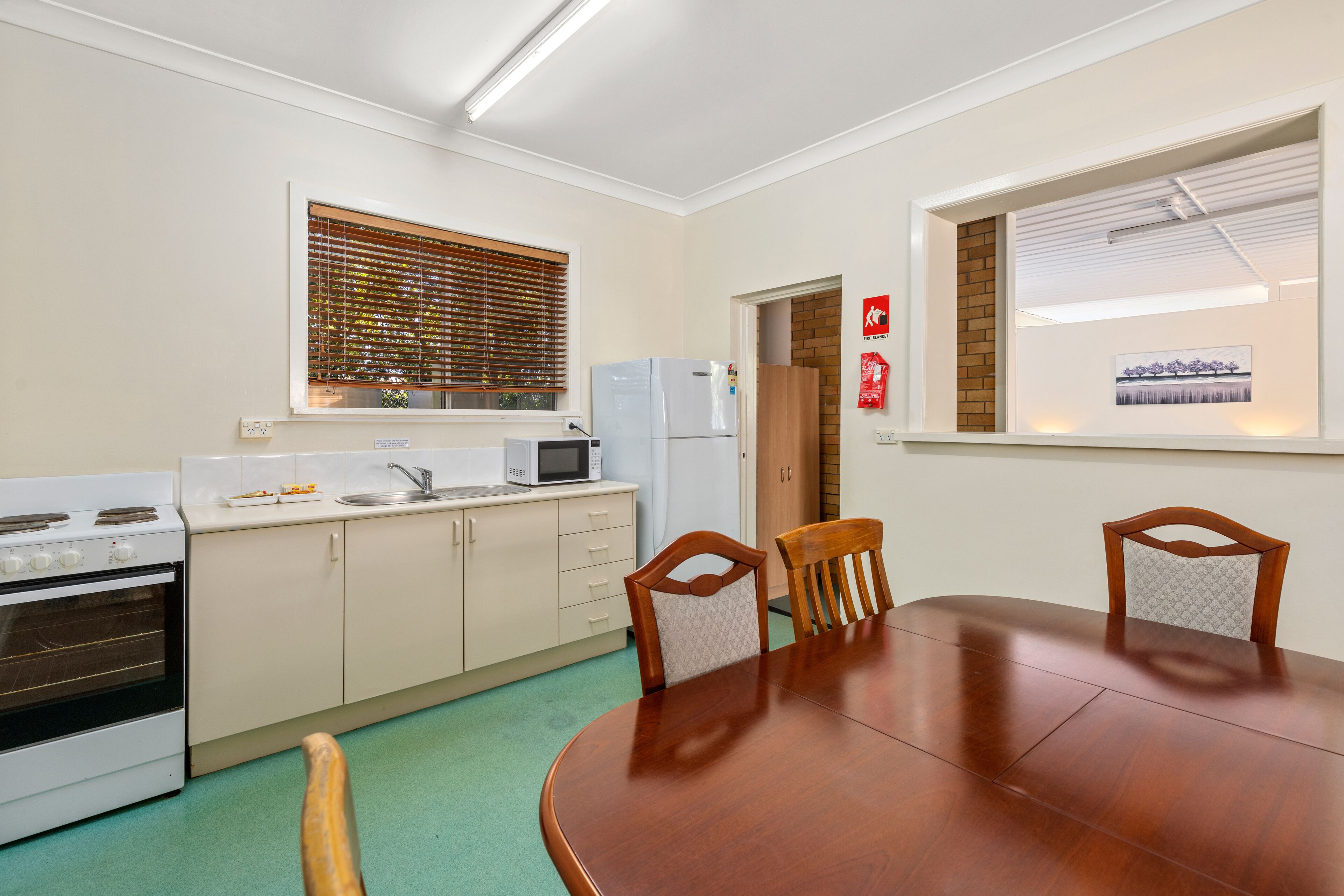 A compact kitchen with a dining table, wooden chairs, a microwave, a refrigerator, and a window with blinds.