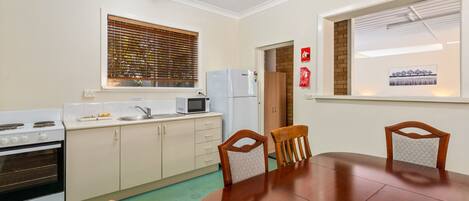 A compact kitchen with a dining table, wooden chairs, a microwave, a refrigerator, and a window with blinds.