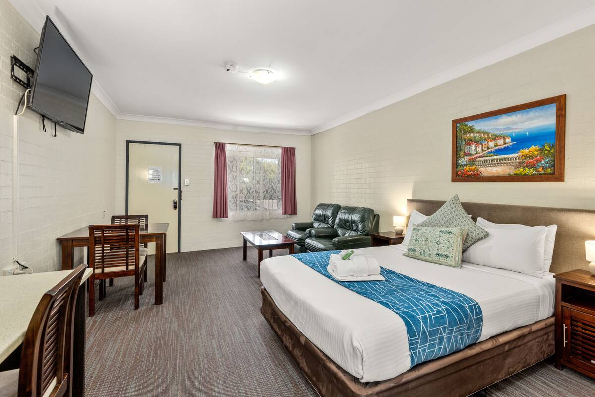 A hotel room with a bed, desk, chairs, a television, and a painting on the wall.