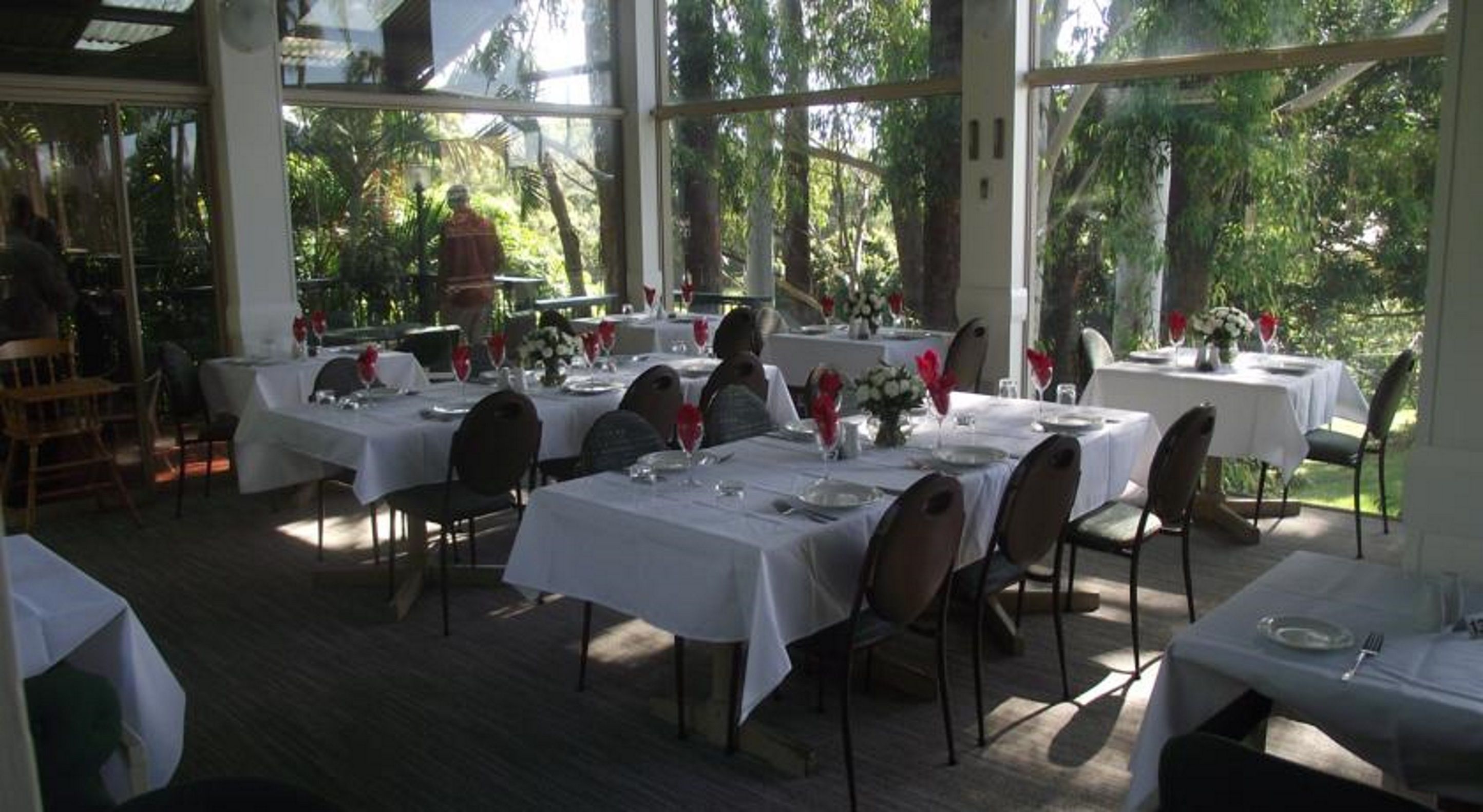 Un espace de restauration en plein air, avec des tables dressées pour le repas, entouré de murs de verre et d’arbres.