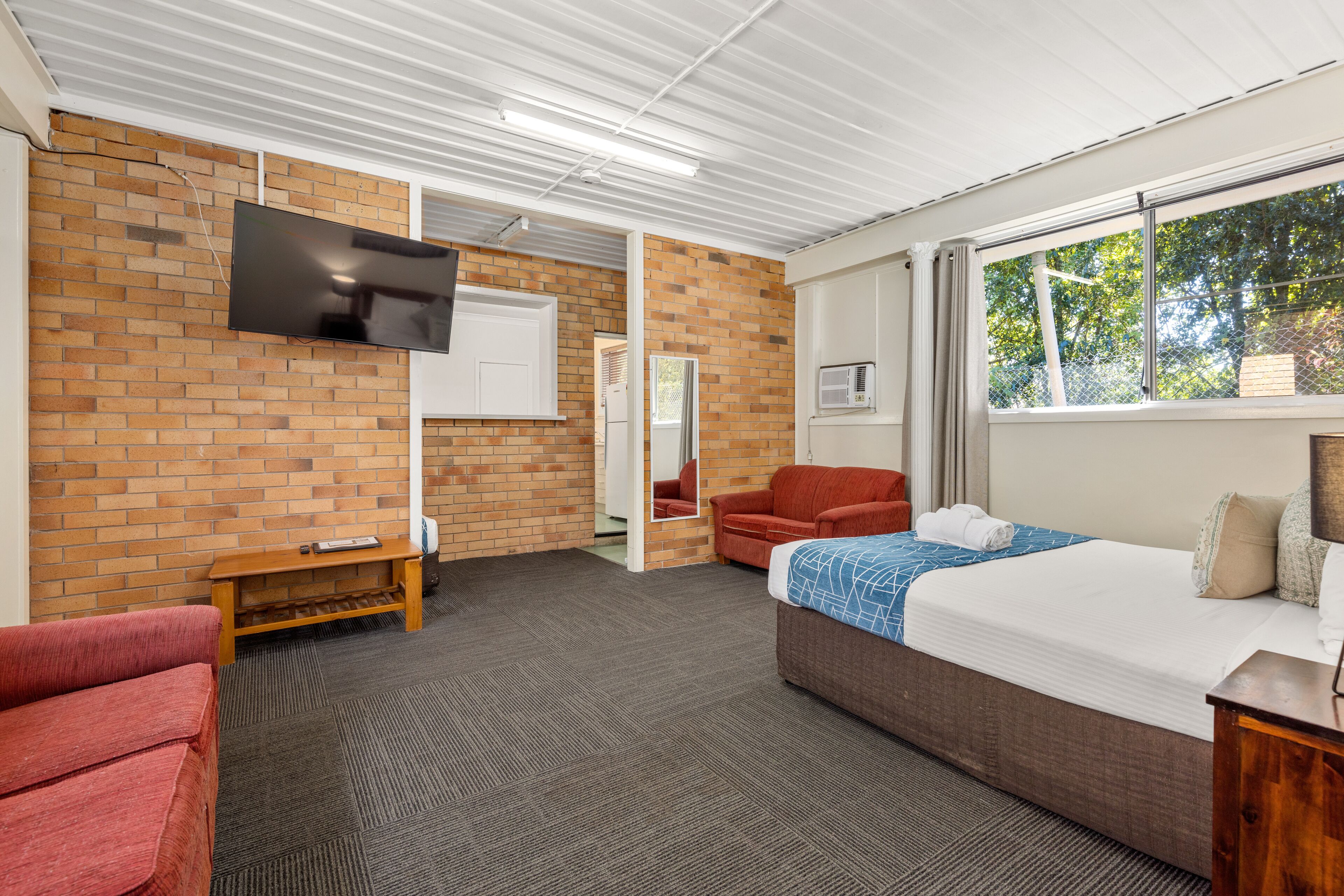 A hotel room with a brick wall, a flat-screen TV, a bed with a blue and white patterned bedspread, a red armchair, and a wooden bench.