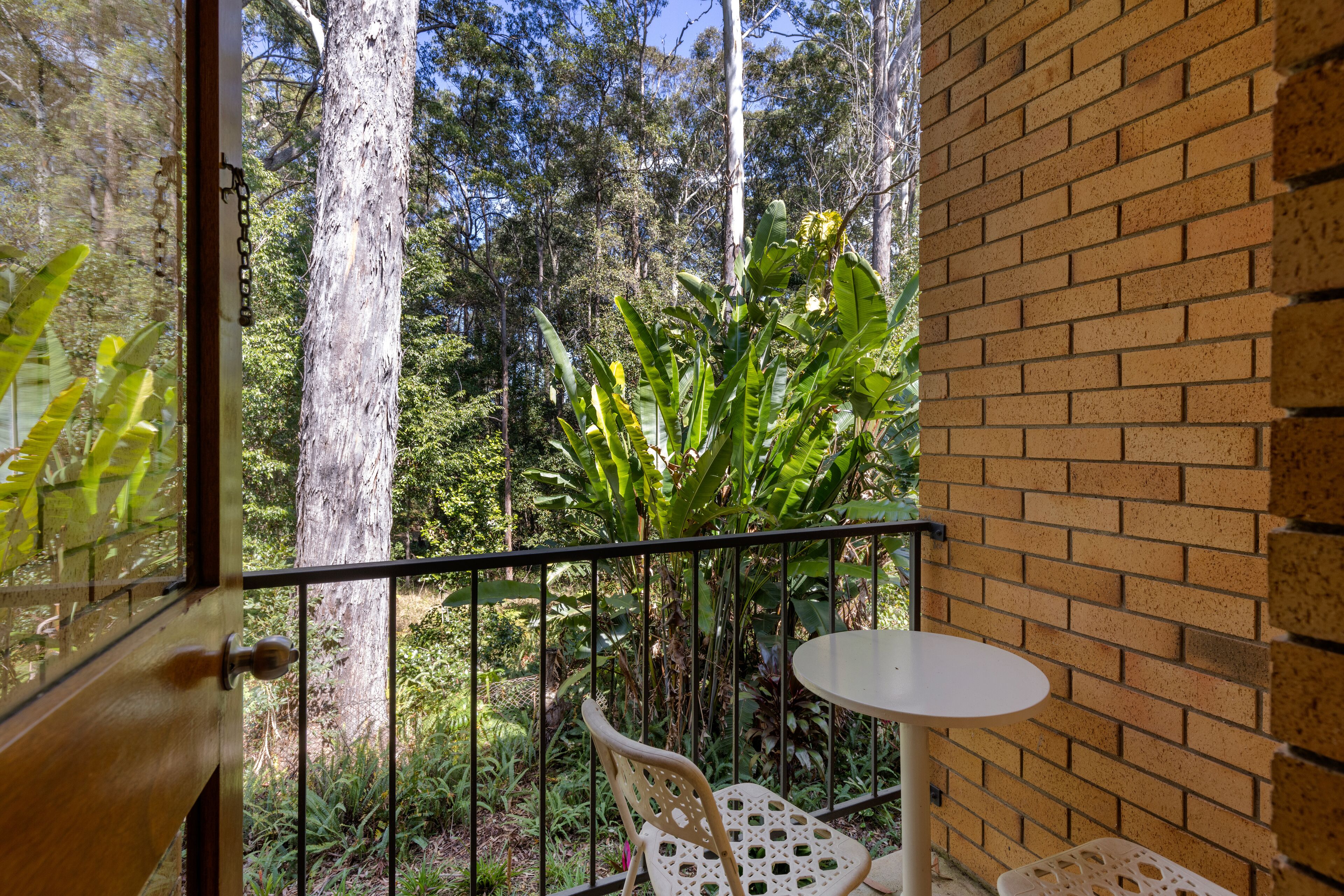En balkon med udsigt over en skov, en murstensvæg og et lille bord med stole.