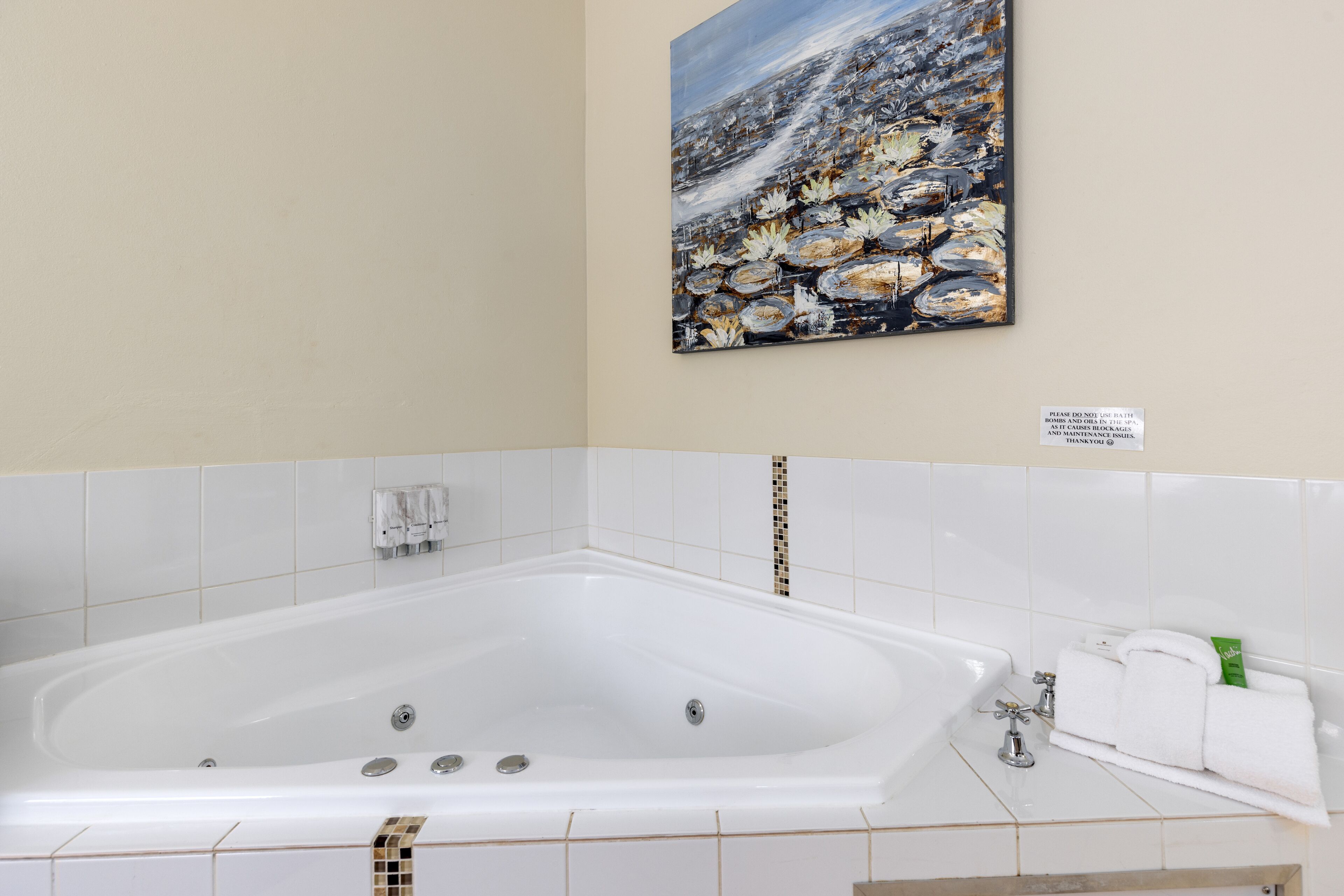 A bathroom with a large bathtub, white tiled walls, and a painting of a coastal scene.