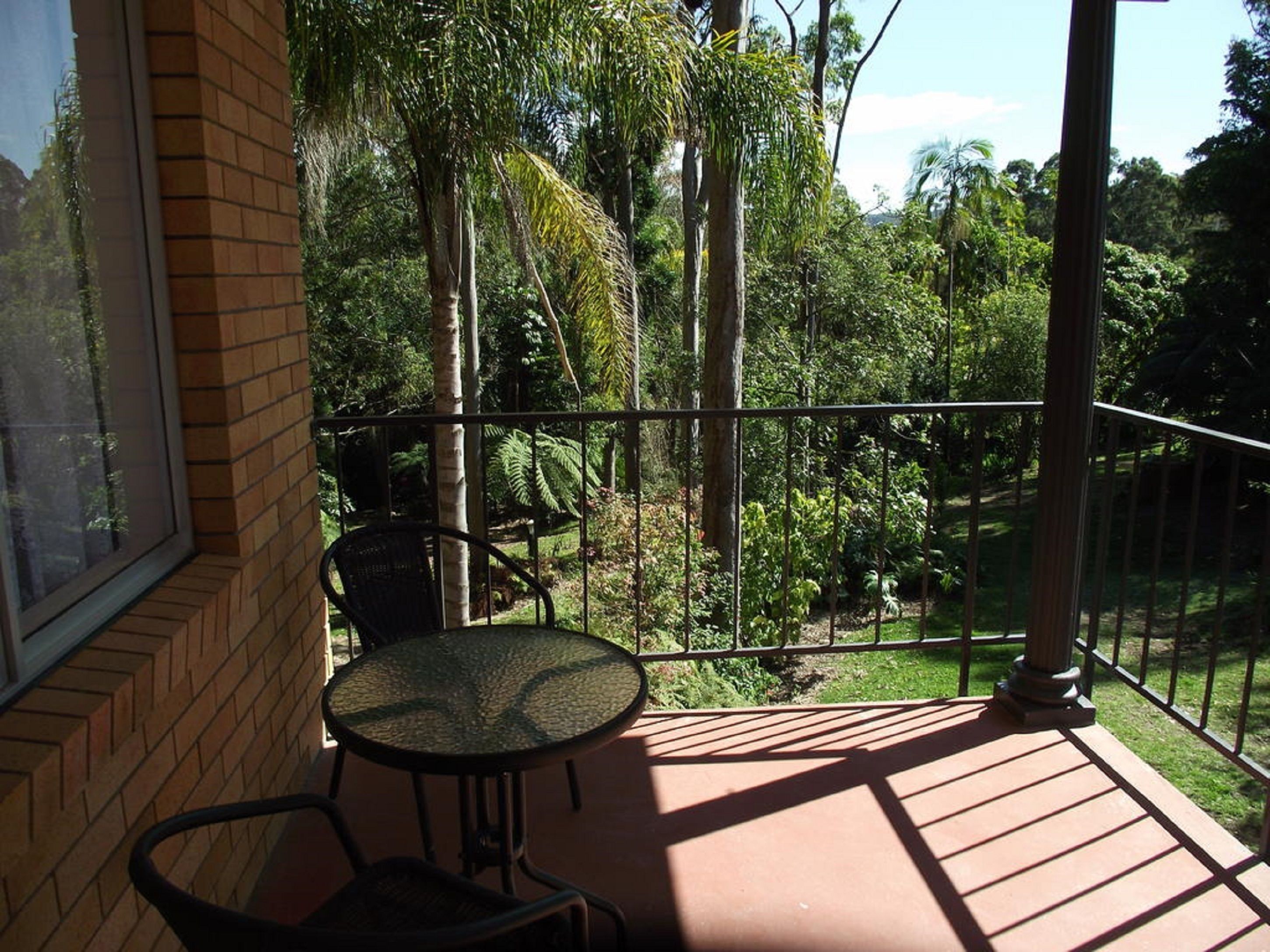 A balcony with a round table and two chairs, overlooking a lush garden with various trees and plants.