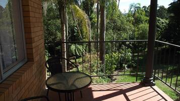 A balcony with a round table and two chairs, overlooking a lush garden with various trees and plants.