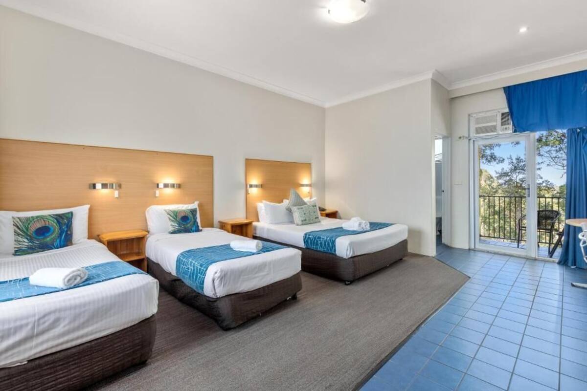A hotel room with three beds, each with a wooden headboard and decorative pillows. There are wall-mounted lights and a large window with a view of the outdoors.