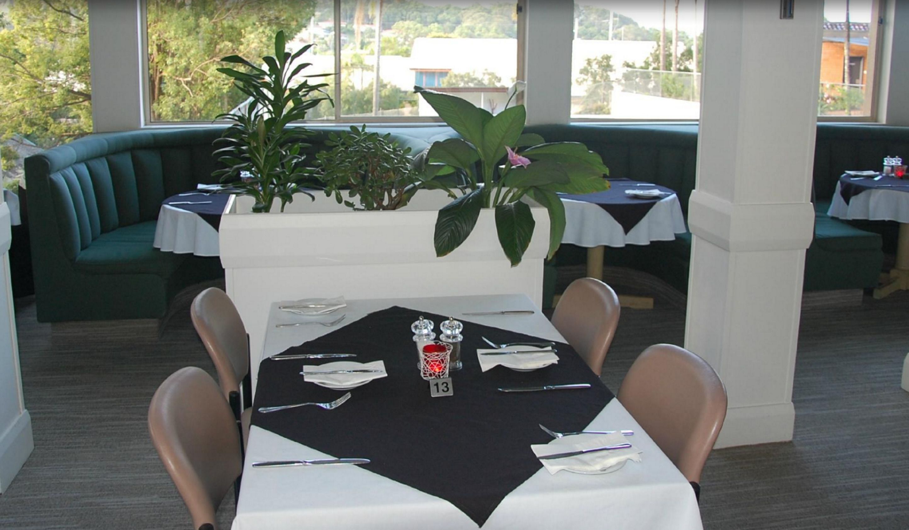 Un espace repas avec une table dressée pour quatre personnes, entouré de banquettes recouvertes d’un tissu vert, et de grandes fenêtres offrant une vue sur l’extérieur.