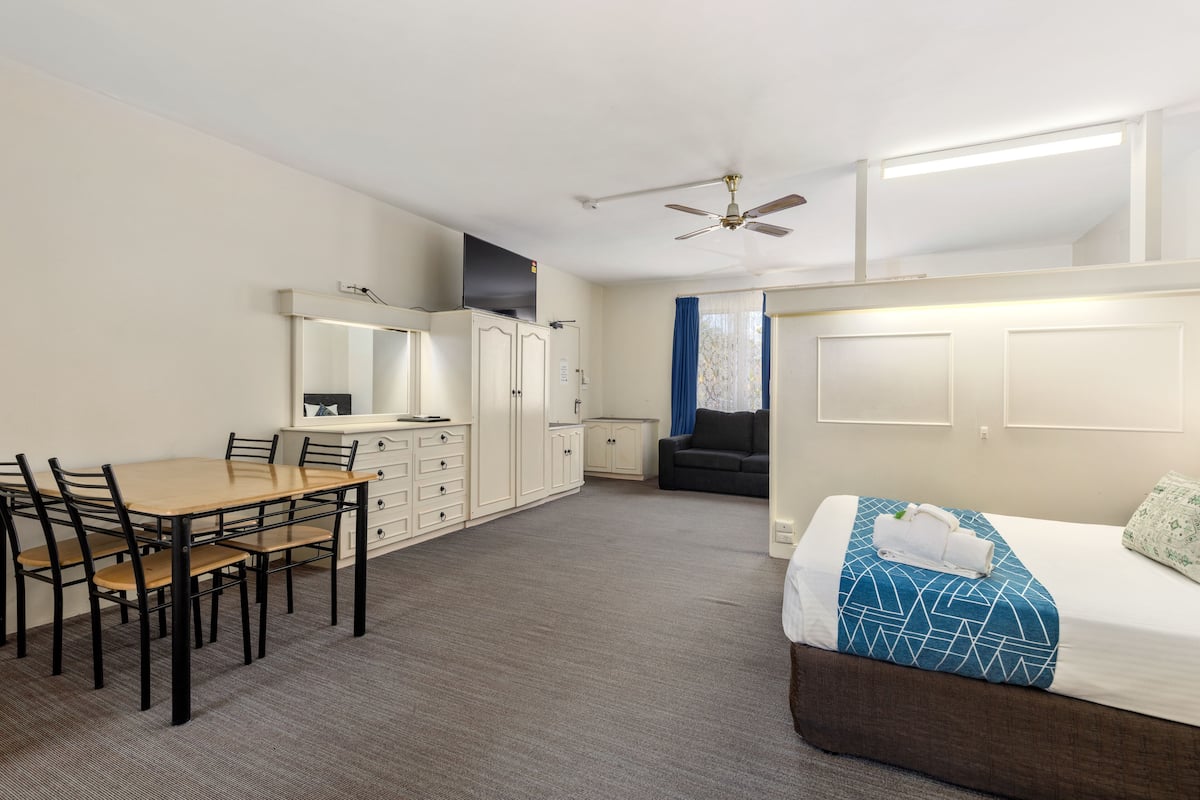 A room with a bed, a desk, and a chair. There is a ceiling fan and a window with curtains.