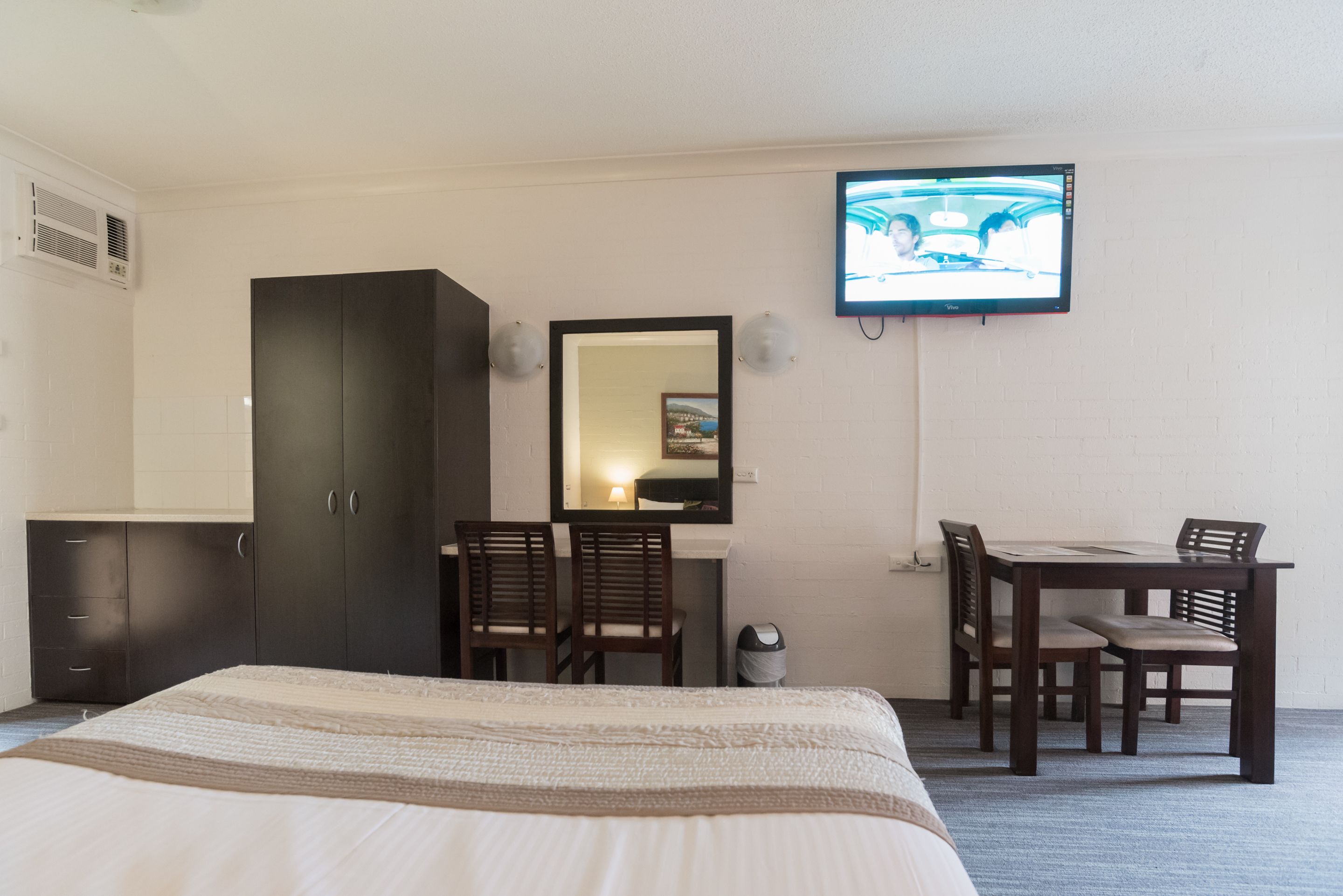 A hotel room with a bed, a desk with two chairs, a TV mounted on the wall, and a wardrobe.