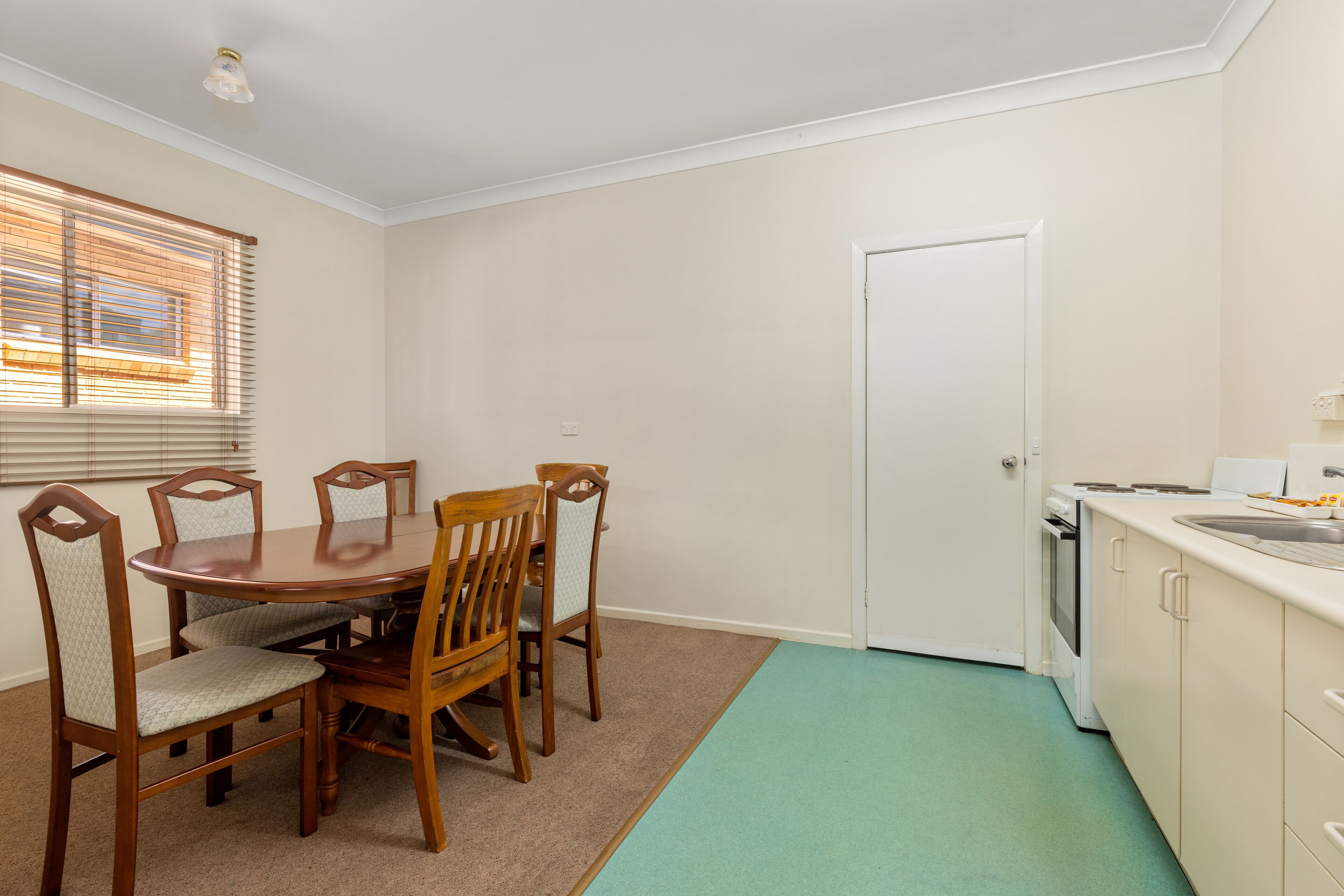 A dining area with a round table and four chairs, a window with blinds, a white door, and a kitchen with a sink and stove.