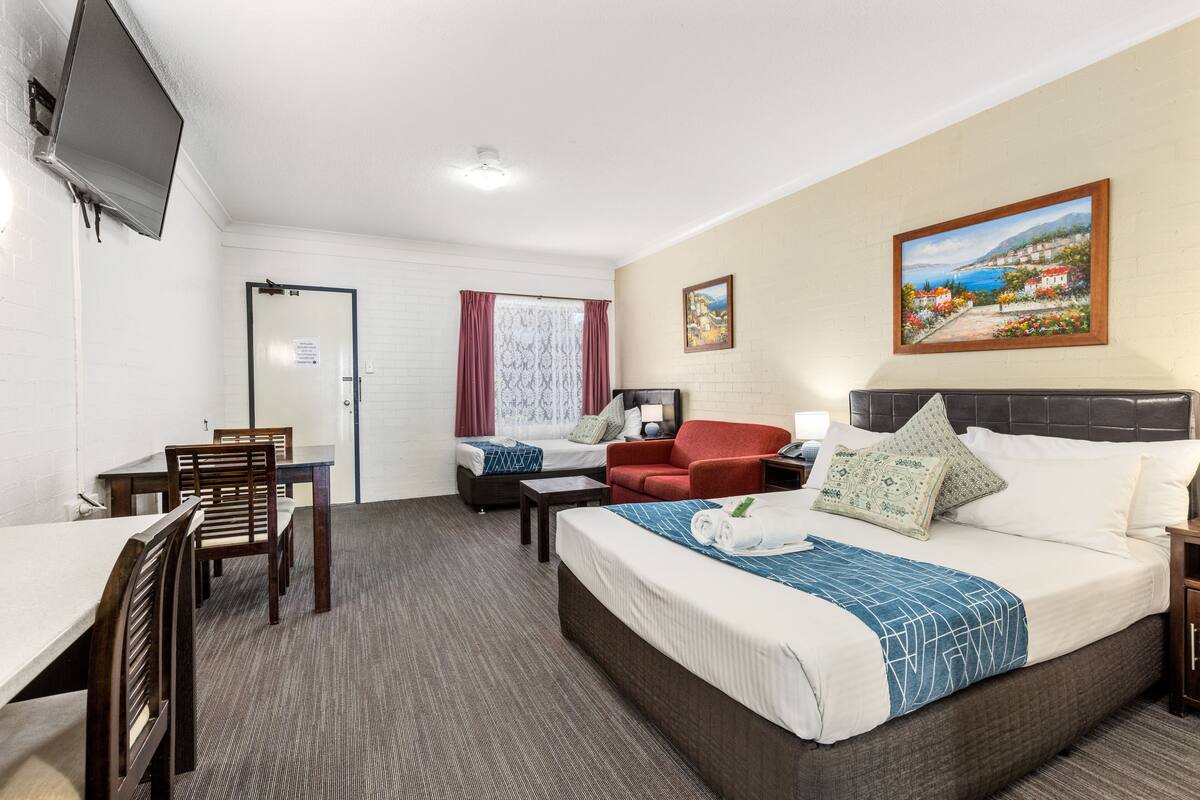 A hotel room with a bed, a desk, a chair, a red sofa, and a painting on the wall.