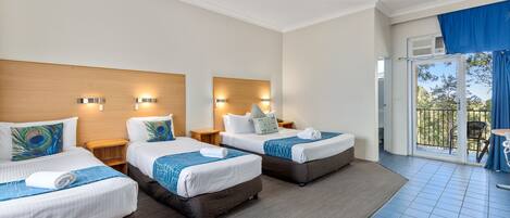 A hotel room with three beds, each with a wooden headboard, white bedding, and decorative pillows. There are wall-mounted lights and a large window with a view of the outdoors.