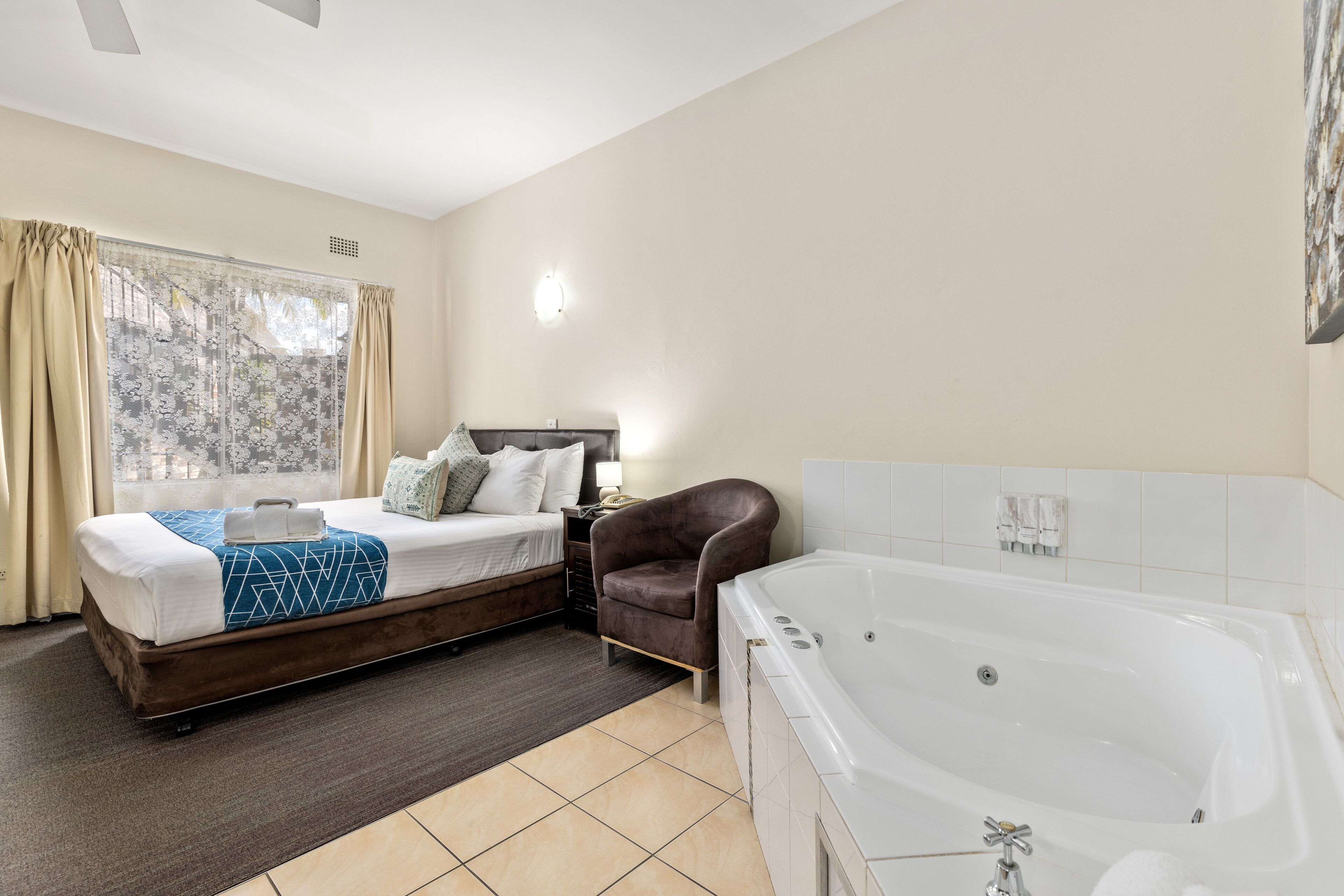 A hotel room with a double bed, a sitting area with a chair, a large bathtub, and a window with curtains.