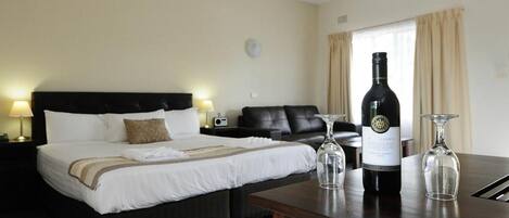 A hotel room with a bed, a sofa, a wooden table with a wine bottle and glasses, and a ceiling fan.