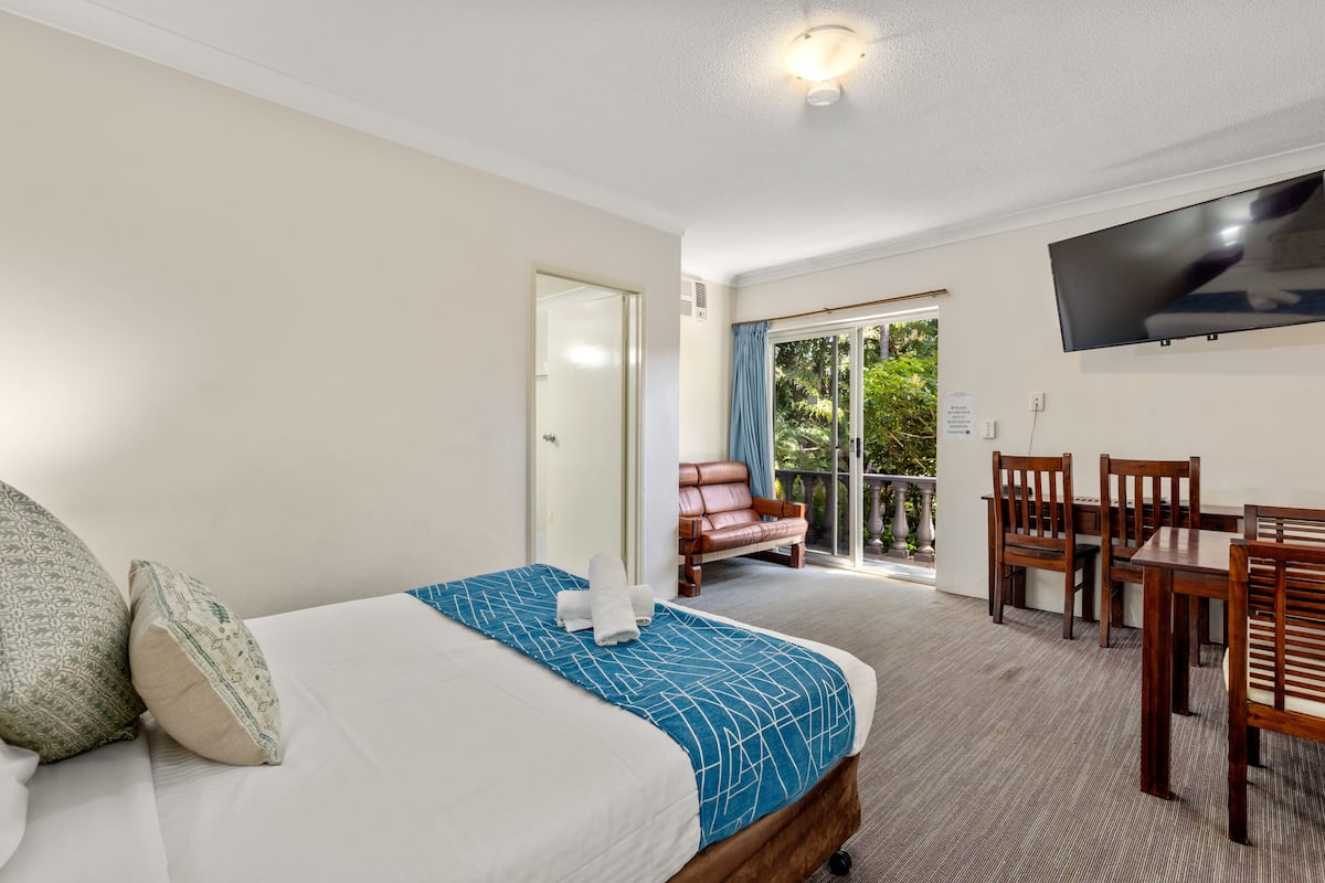 A hotel room with a bed, a dining table with chairs, a TV, and a sliding glass door leading to an outdoor area.