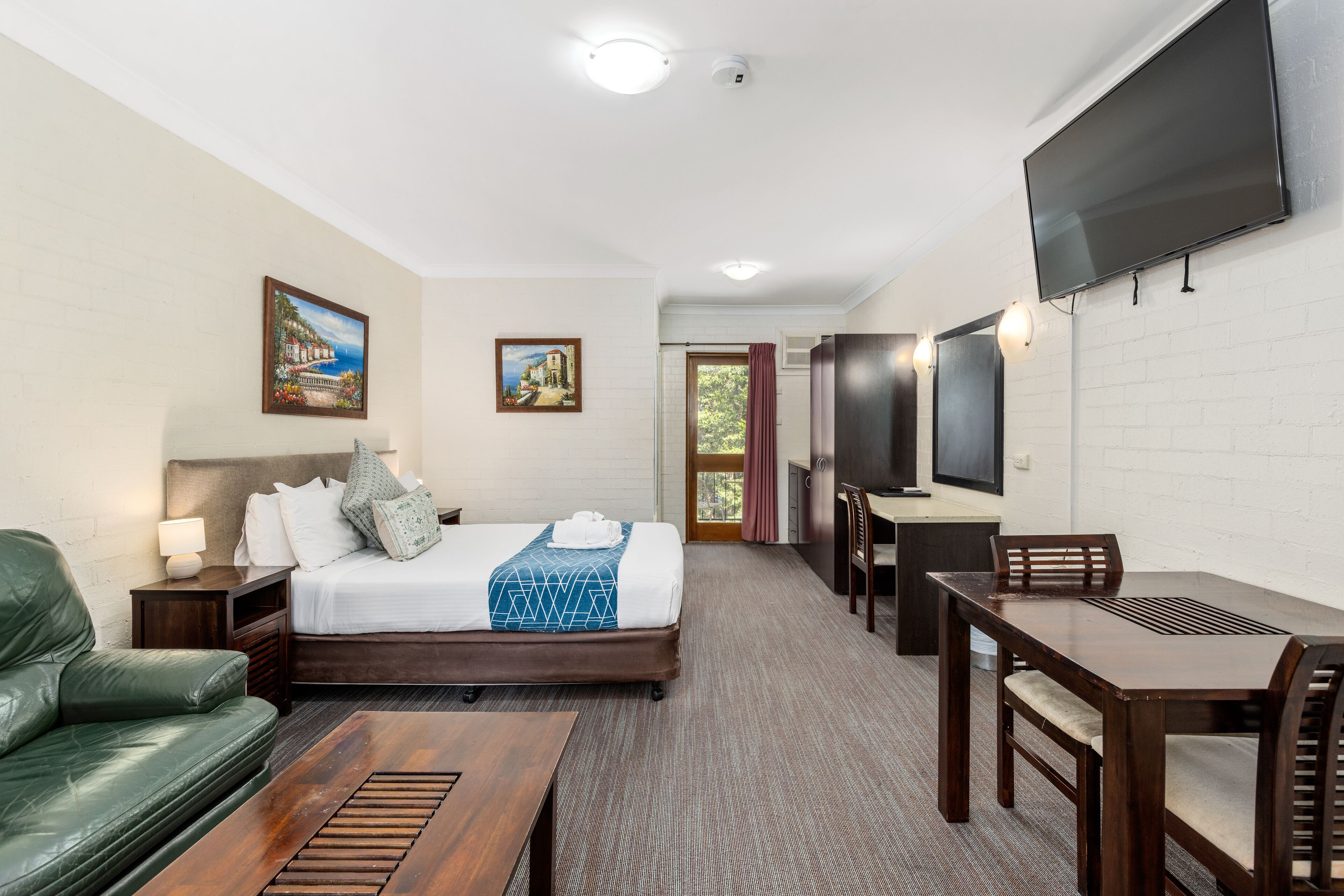 A hotel room with a bed, a green sofa, a wooden table, a TV, and a painting on the wall.