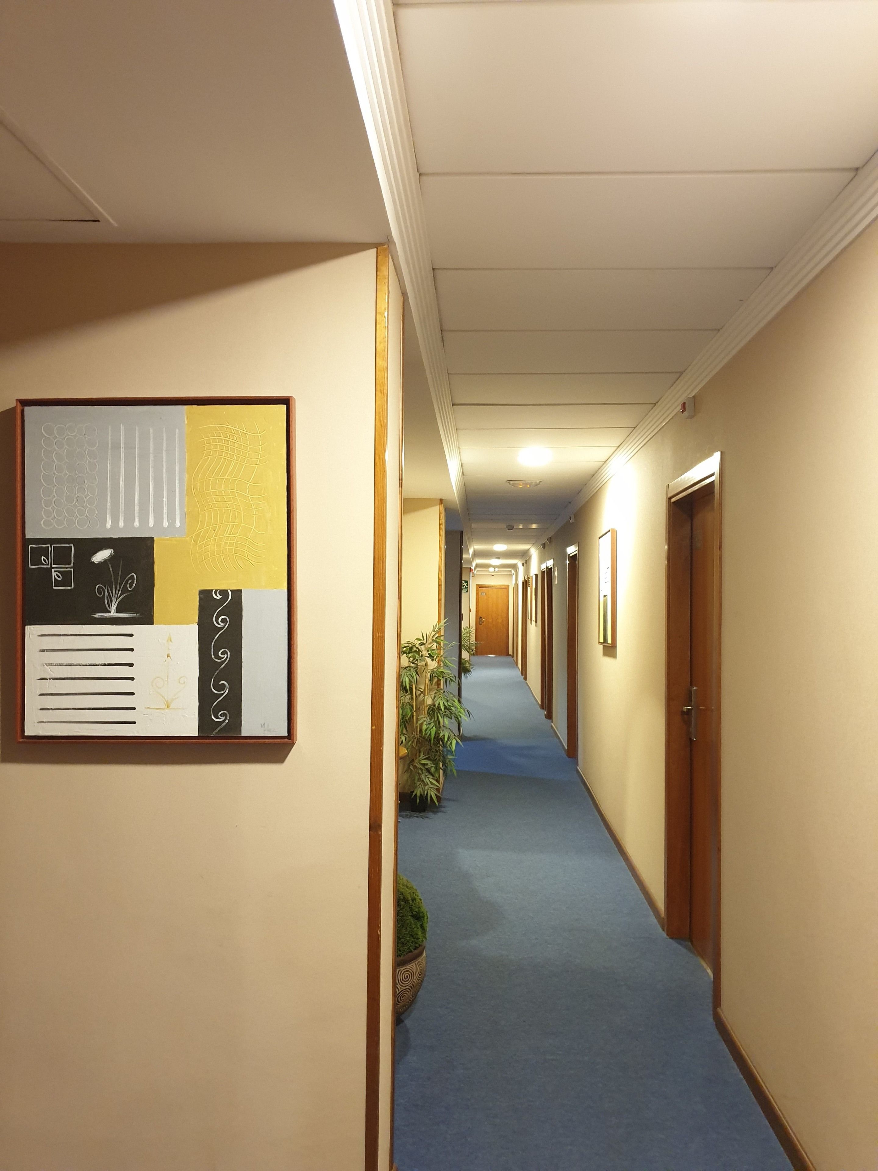 Un corridoio d'albergo con tappeto blu, porte in legno e una bacheca sulla parete sinistra.