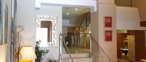 A spacious lobby with a staircase leading to an upper floor, a seating area with a sofa and a table, and a potted plant.