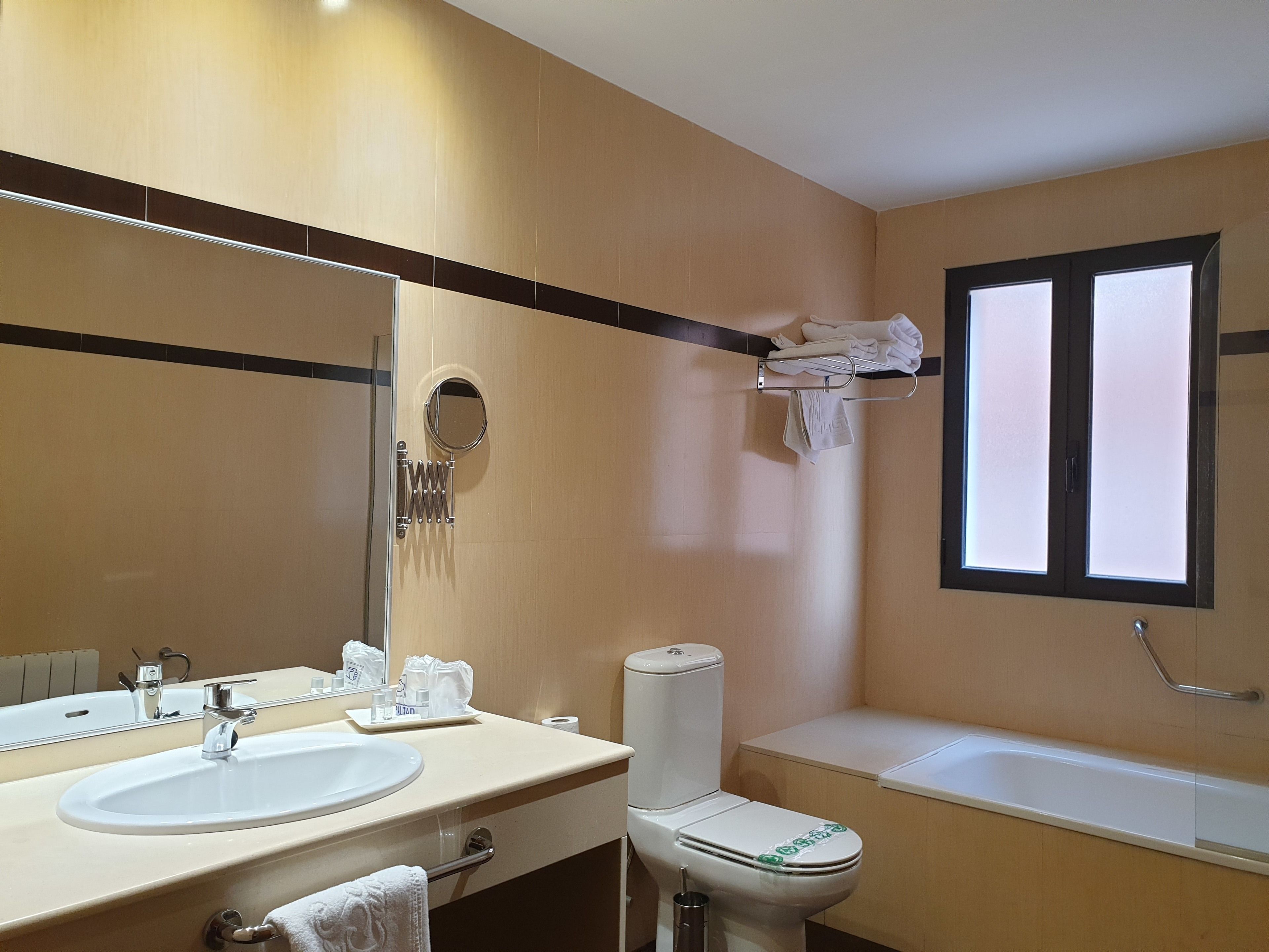 Un baño con lavamanos, inodoro y bañera. Hay un perchero con toallas, un espejo y una ventana.