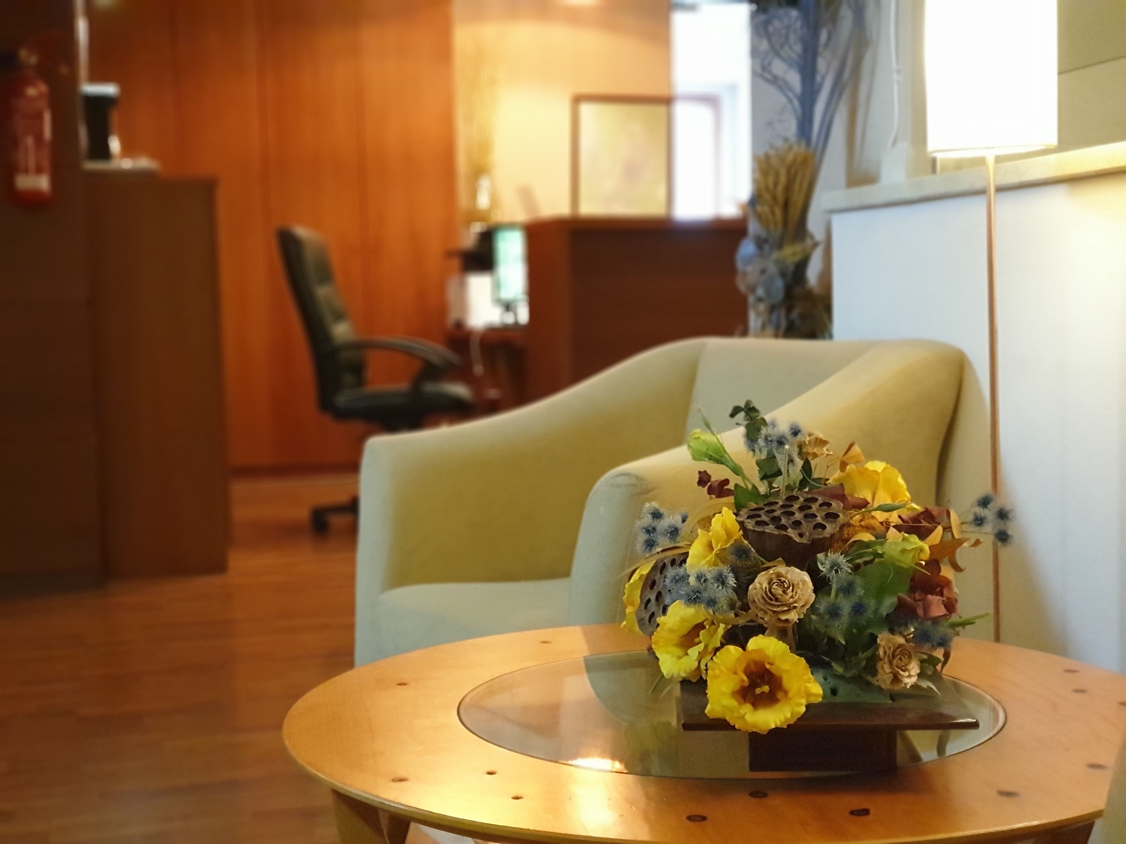 Ein runder Glastisch mit floralem Mittelstück, ein gelber Ohrensessel und ein Holzbüro in einem wohlig beleuchteten Raum mit Holzwänden.