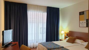 A hotel room with a bed, a desk with a television and remote, a lamp, and a window with curtains.