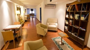 Habitación de hotel con piso de madera, un sillón color beige, una mesita con una lámpara, una estantería y vista al comedor.